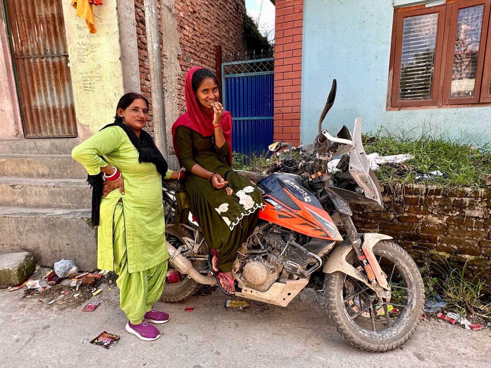 Due donne nepalesi sorridenti accanto alla moto di Antonio Rocco Cossa, davanti a una casa di villaggio in Nepal