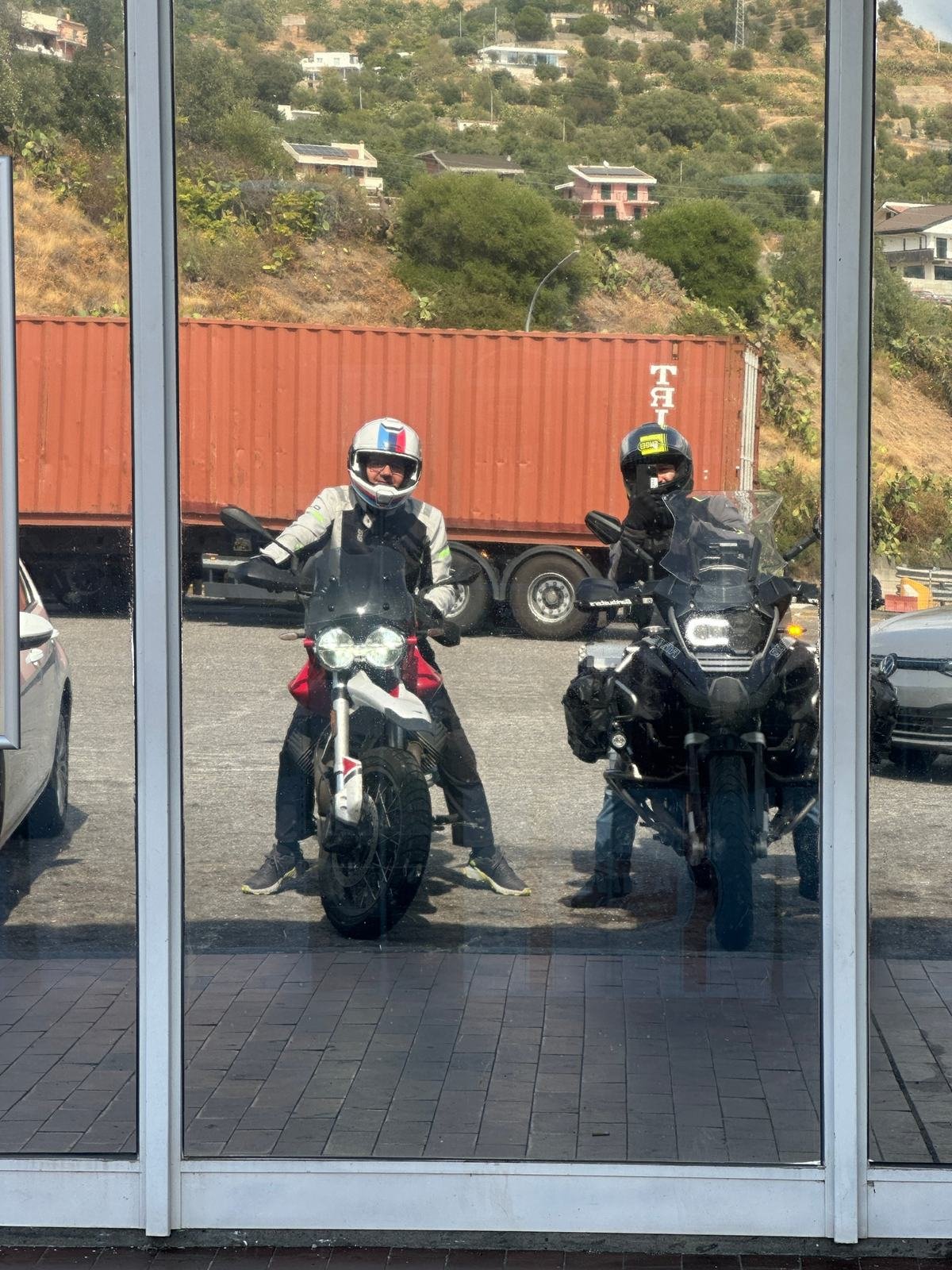 Due motociclisti di Epic Routes Community in sella alle loro moto, riflessi nella vetrata di un'area di servizio sulla strada per la Sicilia